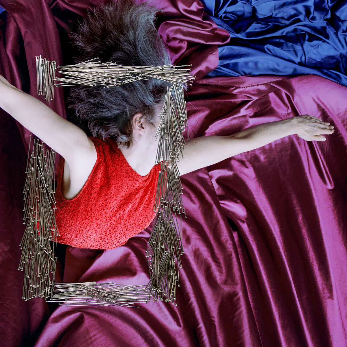 Artwork created with a colour photographic print of a female figure in a bright red dress, set against a purple and blue draped silk background. The figure is viewed as if from above her looking down. Her arms are extended out to the side, hands stretched out. Her body is covered by groups of dress pins, laid on top of the photographic print. The pins are arranged in a square frame shape in the centre of the print. One side of this frame covers her face. Another side of the pin frame stops above and below her extended arm making it look as if she is bursting through the frame.