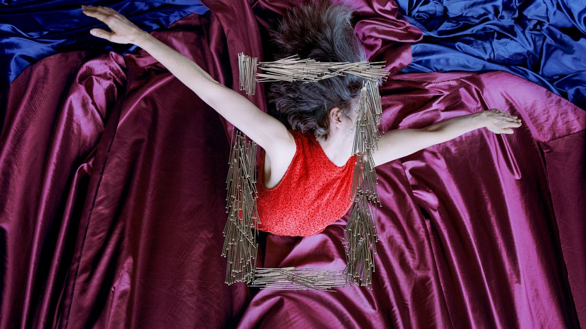 Artwork created with a colour photographic print of a female figure in a bright red dress, set against a purple and blue draped silk background. The figure is viewed as if from above her looking down. Her arms are extended out to the side, hands stretched out. Her body is covered by groups of dress pins, laid on top of the photographic print. The pins are arranged in a square frame shape in the centre of the print. One side of this frame covers her face. Another side of the pin frame stops above and below her extended arm making it look as if she is bursting through the frame.