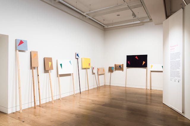 Photograph of an exhibition gallery space showing white walls and a wooden floor. Leaning against the left side wall are several placards on long wooden sticks. Painted on the placards are different coloured triangles. The placards together make the triangles look like bunting strung between the placards. On the wall to the right is a large information text panel.