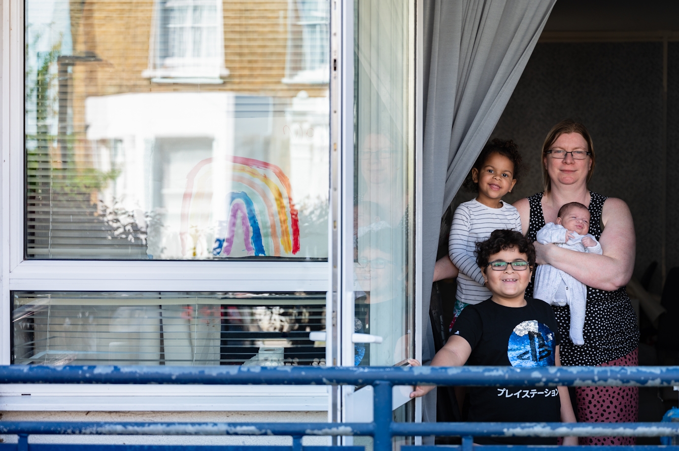Photograph of a mother and her 3 children stood in the open door of their home, smiling to camera. In their window to the left is a hand painted picture of a rainbow.