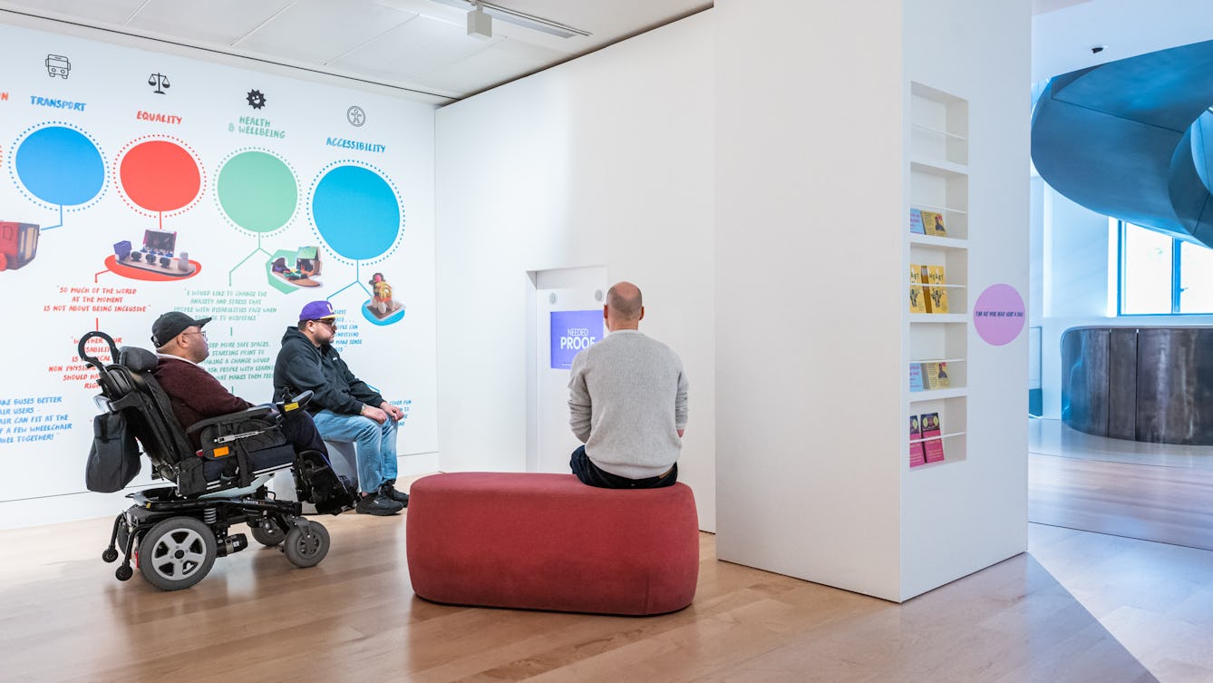 Photograph of a gallery installation with three seated people watching a video on a tv screen. On the wall to the left are large bold graphics and text, coloured blue, red and green. To the right are leaflets standing on 5 shelves, inset into a wall. In the distance are the windows and a section of a spiral staircase.