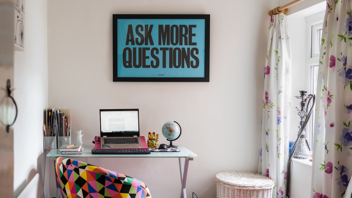 Photograph of a room in a house showing a small desk area with a laptop open on a stand. Next to the laptop is a small globe, note books and file tidies. In front of the desk is a colourful triangular patterned material covered chair. To the right of the desk is a wicker laundry bin, a window with floral curtains and a shisha  on the window sill. Hung on the wall above the desk is a large framed picture with a blue background and large black capital letters with the words, 'ASK MORE QUESTIONS'.