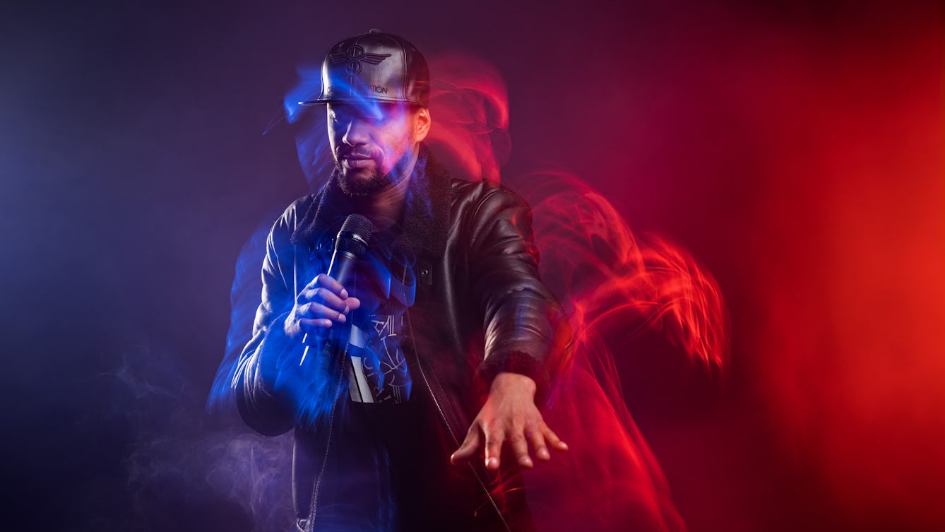 A male rap artist dressed in a black leather hat and jacket is holding a microphone, dancing with his arm outstretched in front of him. The room is filled with smoke and blue and red lighting effects highlight his movement. 