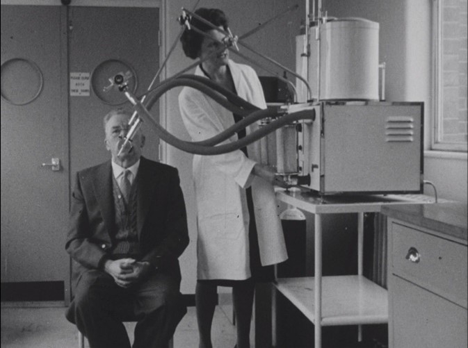 Black and white film-still showing a woman with dark hair in a white doctor's coat operating box-like machinery with tubes running towards a suited man, who is breathing into a mask attached to the tubes. 