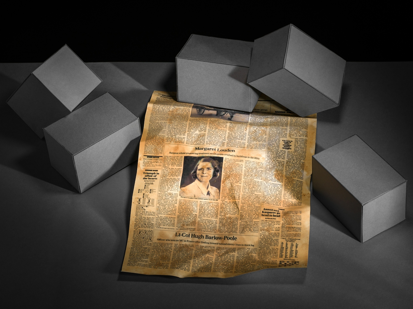 Photograph of a set-built scene made up of a grey card horizontal base against a black vertical background. The view looks down on the surface where there are five rectangular brick blocks scattered in a random arrangement which are also made out of grey cardboard. In the centre of the image and tucked under a couple of the blocks, is an obituary page from a newspaper. It is crumpled and yellowed with age. In the centre of the newspaper is an obituary with a portrait of a young woman with short hair wearing a blouse. Above the portrait is the name Margaret Louden.