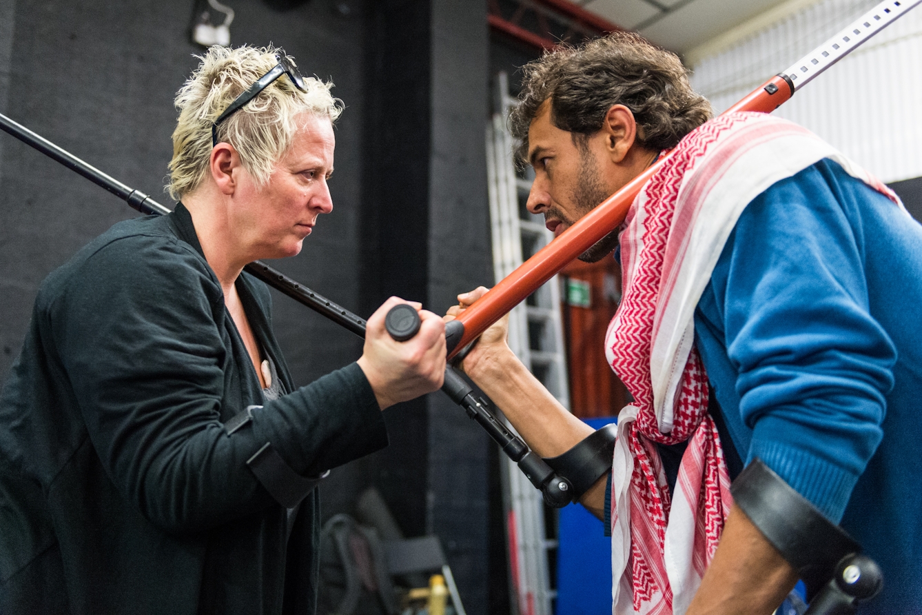 Photograph of a man and a woman in a rehearsal space facing each other in a theatrical expression of competitiveness. Each person is holding a crutch over the shoulder of the other.  The arms and crutch of each person overlap and form an X at the centre of the frame.