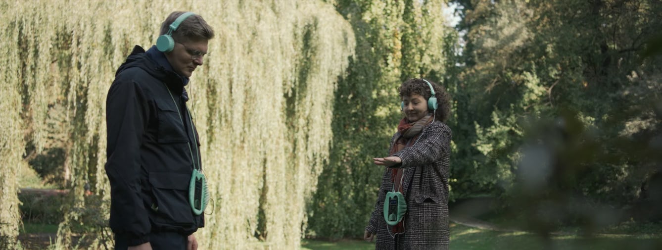 Two people in a park wearing over ear-headphones connected to digital devices which are hanging around their neck. They are both looking down listening to their headphones. One person is holding their hand out.  