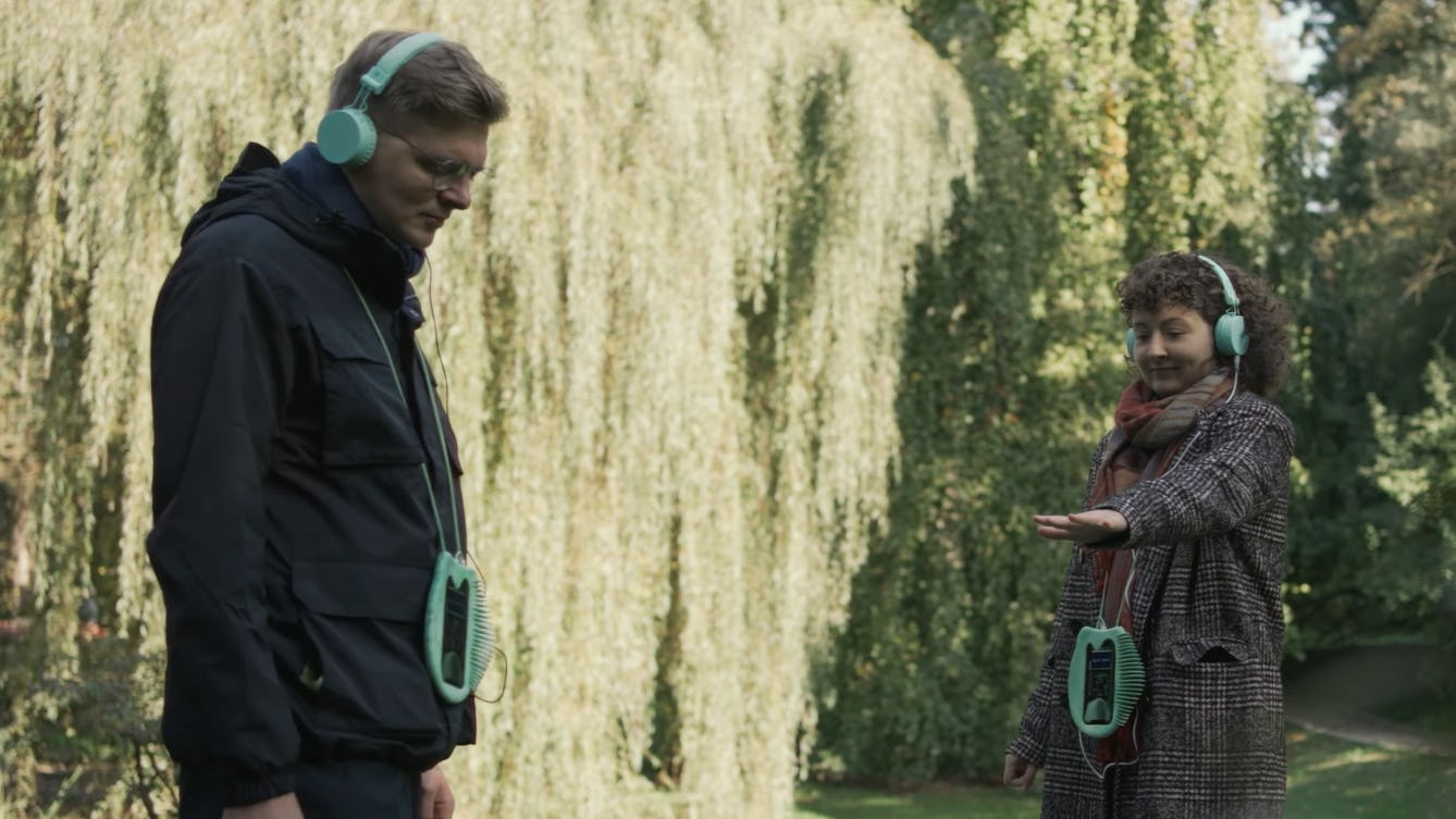 Two people in a park wearing over ear-headphones connected to digital devices which are hanging around their neck. They are both looking down listening to their headphones. One person is holding their hand out.  