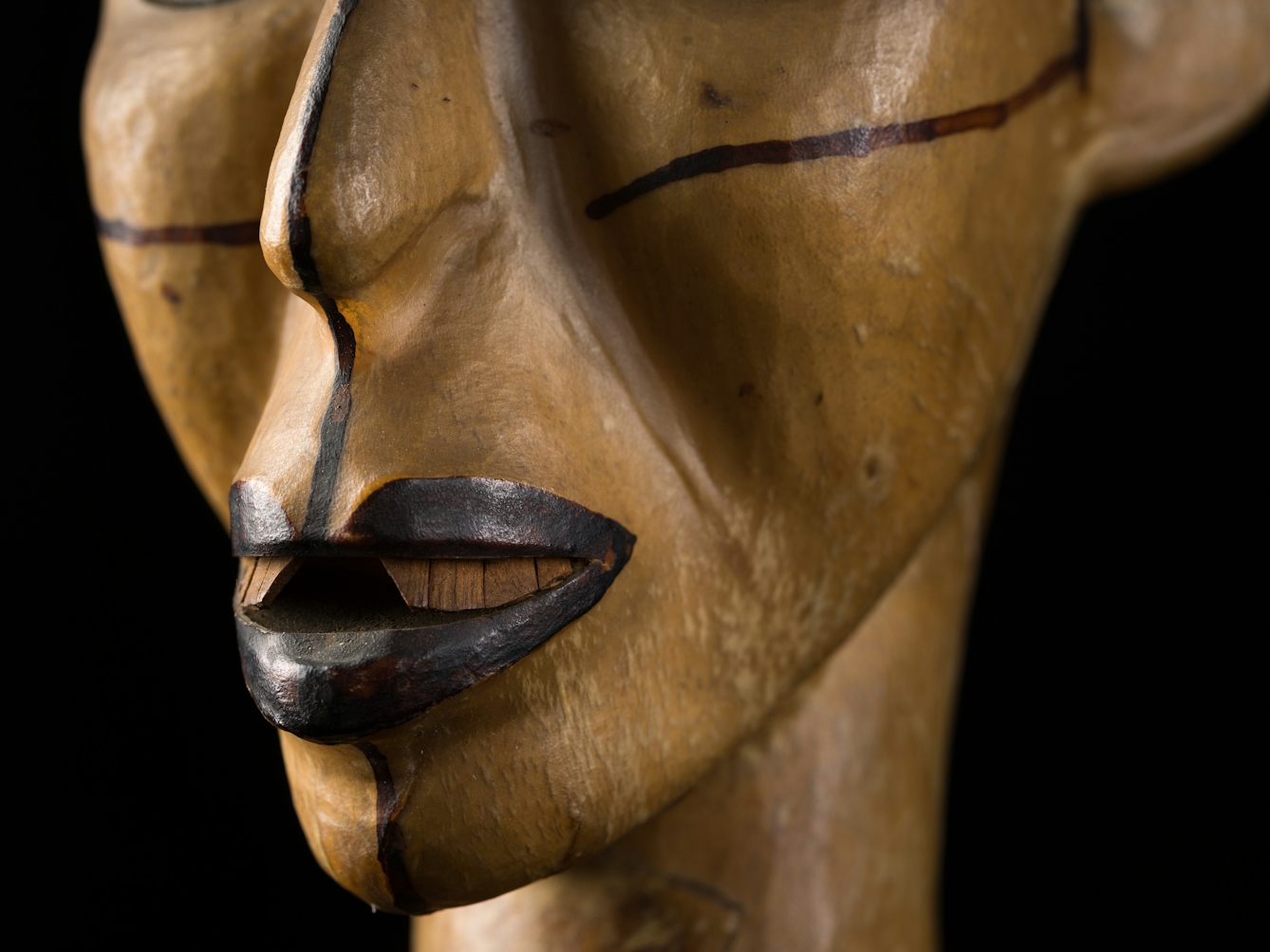 Photograph of the carved wooden head of a woman against a black background. The image shows a close-up detail of her nose and mouth.