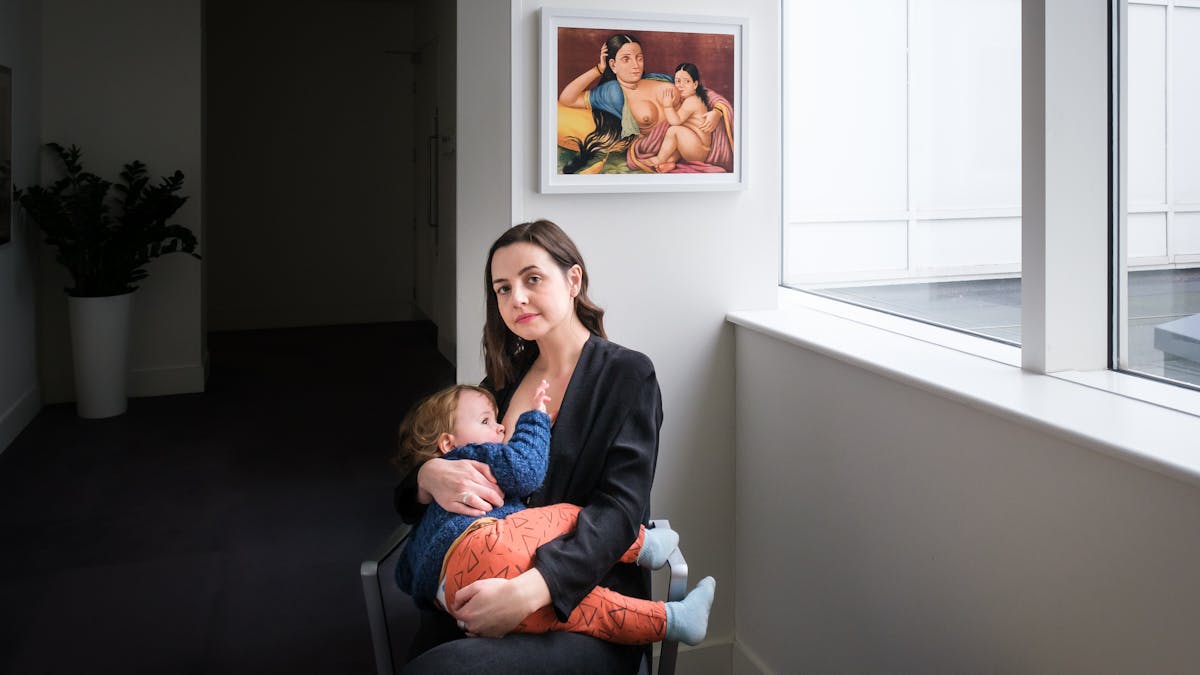 Photograph of a seated woman, who is breasting her son, cradled in her arms. Behind her is a framed painting of a reclining mother and child, also depicted breastfeeding. To the right of the mother is a large window with natural light flooding in. To the left is a corridor in shadows with a leafy plant in a tall white pot in the distance.