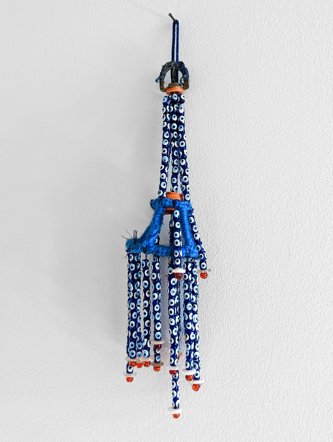 Strands of blue and white glass beads containing the circular 'evil eye' symbol form a wall-hanging, attached to a white wall by a single nail. Teh strands are connected together with reycled bootle tops and wool.
