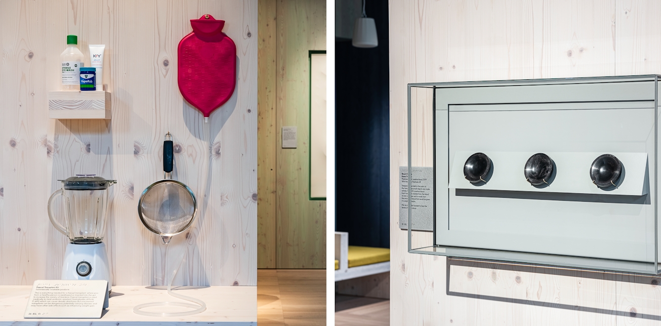 Diptych. The photo on the left shows a wooden panelled table top on which sits a food blender. A wooden panelled surround holds a small shelf on which sterile eye wash, VapoRub, and KY Jelly sit. To the right, hanging from a hook, is what resembles a pink hot water bottle with a valve at the bottom with a long clear plastic tube attached and trailing down the backboard onto the wooden table top. The photo on the right shows a glass case mounted on a wooden panelled wall containing three black marble like palm sized plasticised blood dishes in a line.