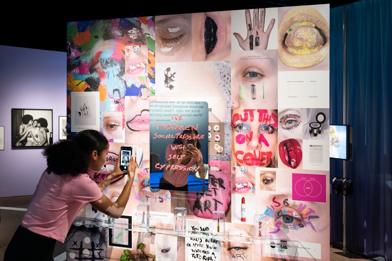 Photograph of a gallery visitor taking a photo of herself with a phone in the mirror at the centre of an installation. The mirror is surrounded by colour photos of lips, eyes and hands, some covered in make-up and hand written words. Scrawled across the mirror in what looks like red lipstick are the words, 'I've mistaken social pressure with self expression'.