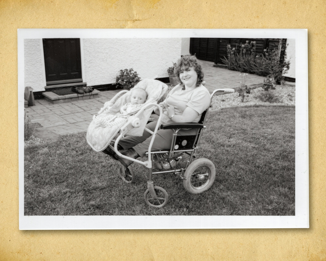 Photograph of a black and white photographic print, resting on a brown paper textured background. The print shows a garden scene with a grass lawn and part of a white rendered house and black door. In the centre of the image is a woman seated in a wheelchair. Her arms and legs are short as a result of her mother being prescribed thalidomide when she was pregnant. Attached to the front of her wheelchair is a baby carrier with a small baby strapped in. Both are looking towards camera, the mother smiling.