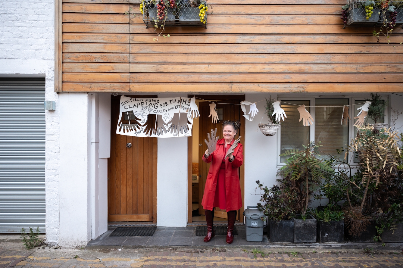 Photograph of the front of a house with wooden cladding and bunting made out of paper cut out hands and a sign that reads, 'Harefield Mews clapping for the NHS carers and key workers'. In the doorway stands a woman in a red coat, holding large cardboard hands in the process of clapping.