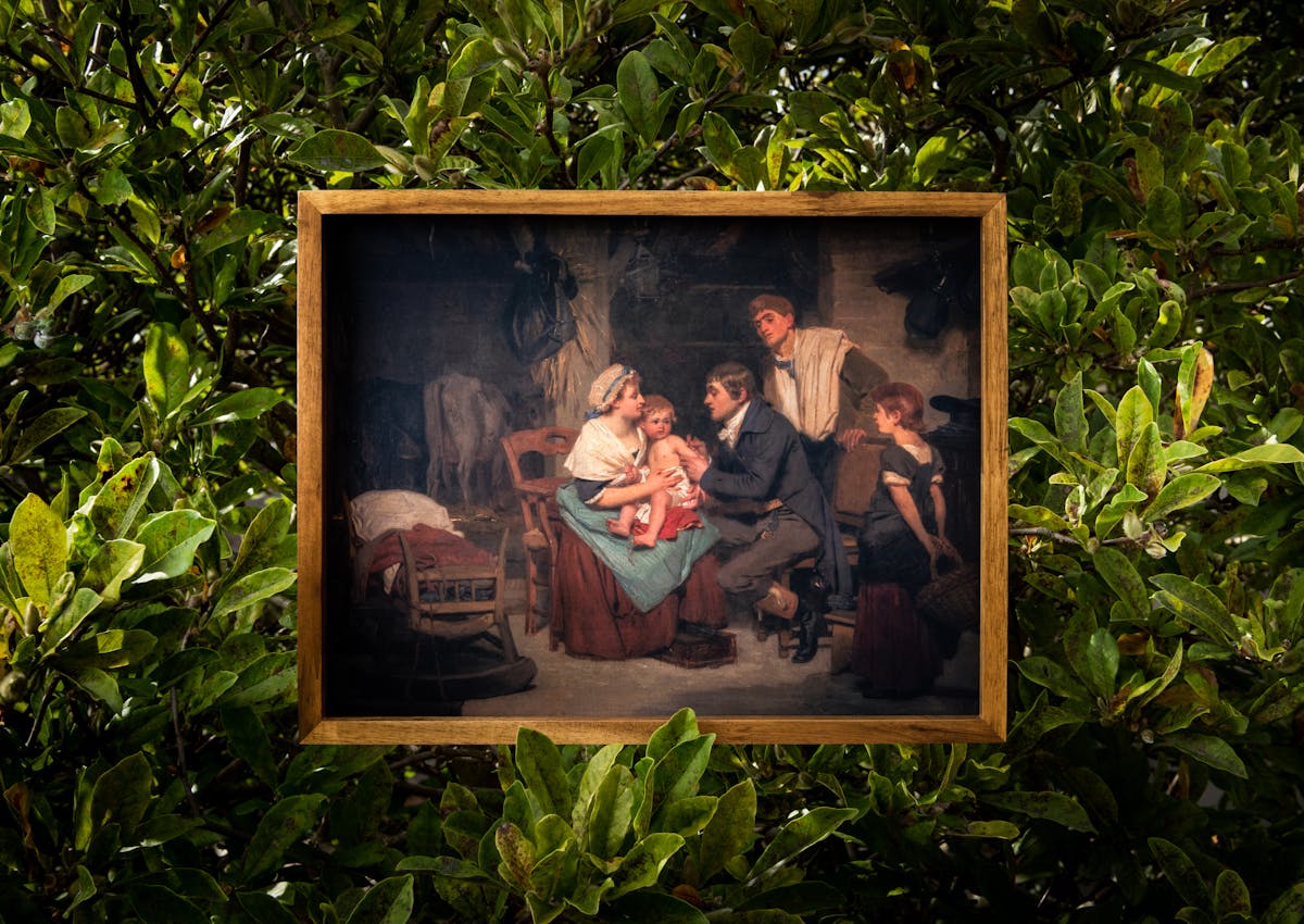 Photograph of a facsimile of an oil painting in a thin wooden frame, hung within the large green leaf foliage of a bush. The oil painting depicts a scene where a man is giving a small child a vaccination. The child is sat on it's mother's knee and the older sister and father figures look on. The scene is set in the 18th century.