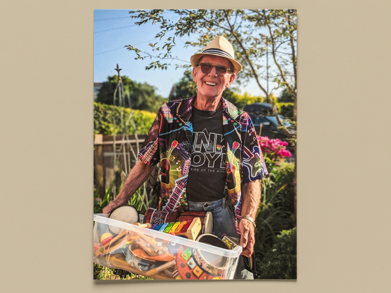 Photograph of a print of a colour photo resting on a light taupe-coloured background. The photo shows an older man standing in a garden scene bathed in the sunshine, smiling to camera. He is wearing sunglasses, a trilby hat, and a brightly coloured, patterned, short-sleeved shirt. He is carrying a plastic storage box full of musical instruments, drums, tambourines, xylophones and shakers.