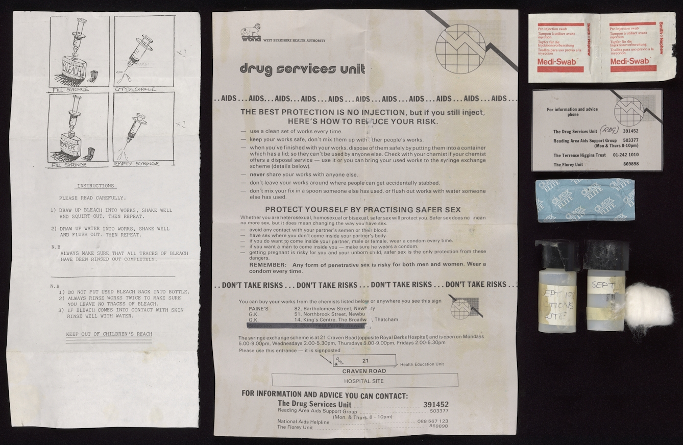 A number of items photographed against a black background, including two black and white leaflets, two medi swabs, a condom, two small screw lid containers, a cotton wool ball and a card with helpful telephone numbers listed on it.