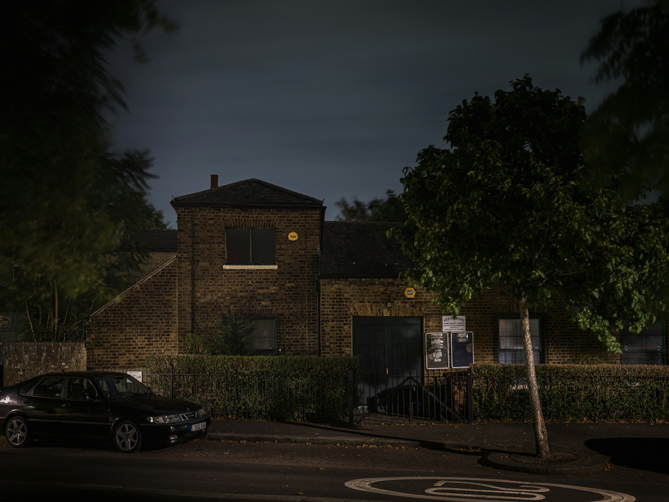 Photograph of the National Spiritualist Church, Walthamstow, at night.  The building is set amongst dense foliage with very little light making its way onto the facade of the exposed brick building.  A single car can be seen in the foreground,