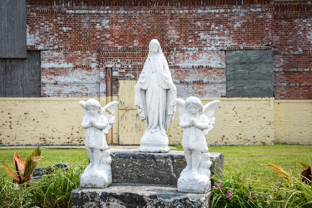 Photograph of three white stone memorial figures, two angels and a robed female. either side of the memorial is long glass and flowers. In the background is a painted brick wall where the paint has flaked off and the doors and windows are boarded up.