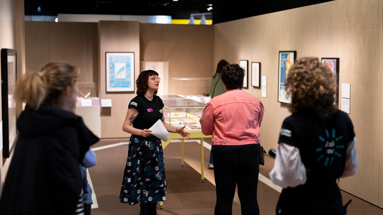 A Visitor Experience Assistant standing in a brightly lit gallery giving a tour of the exhibition to visitors. Two museum staff are wearing black t-shirts with "Wellcome Collection" printed in white lettering on their sleeves. 