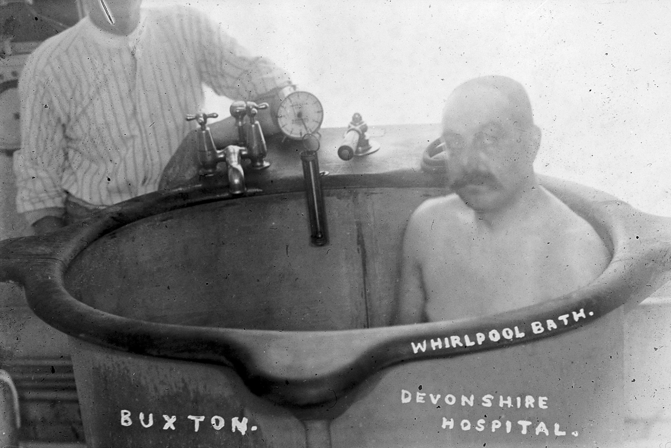 Black and white photograph of a moustachioed man sitting upright in a bath. Behind him stands someone in a blouse leaning on their elbow beside some taps and gauges.