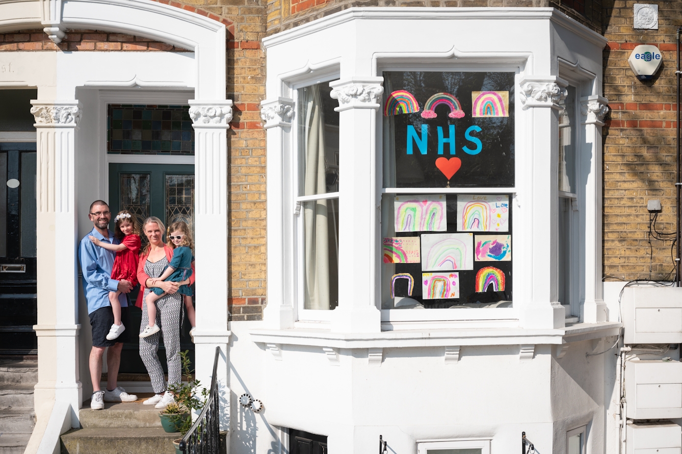 Photograph of the front of a Victoria house showing the front door and bay window. In the doorway stands a father and mother holding their two children. Stuck to the bay windows are hand drawn pictures of rainbows and the letters, NHS with a red heart.