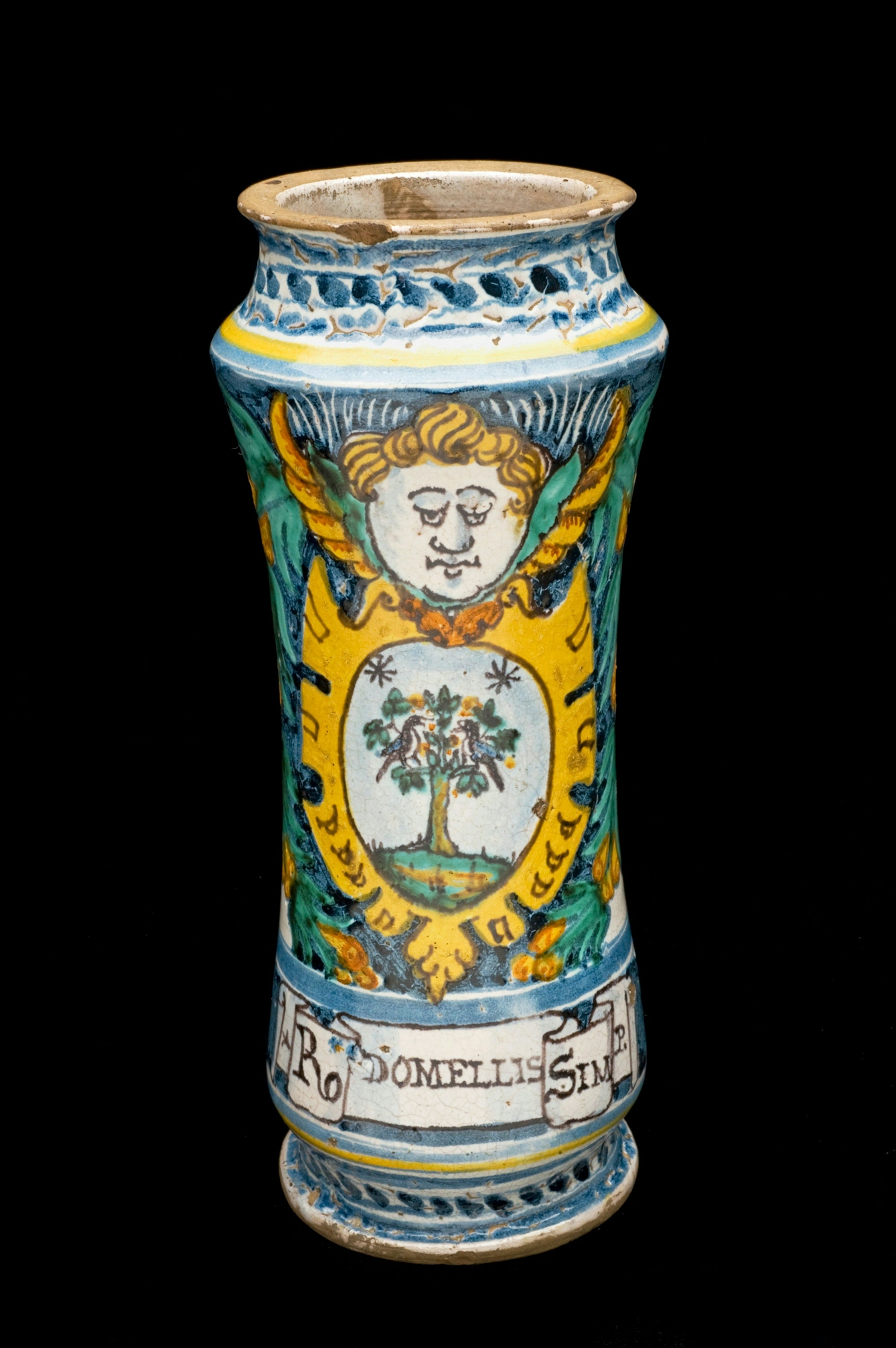 An 18th-century ceramic alberlello drug jar made in Italy. The tall narrow vase-like jar has blue, yellow and green glaze on a white background and depicts a blond cherub face with wings on either side. Beneath this in a yello crest is a picture of two birds in a tree. Bellow this is a scroll with the words "Rhodomel Simplex" (Latin for “Simple Honey of Roses).