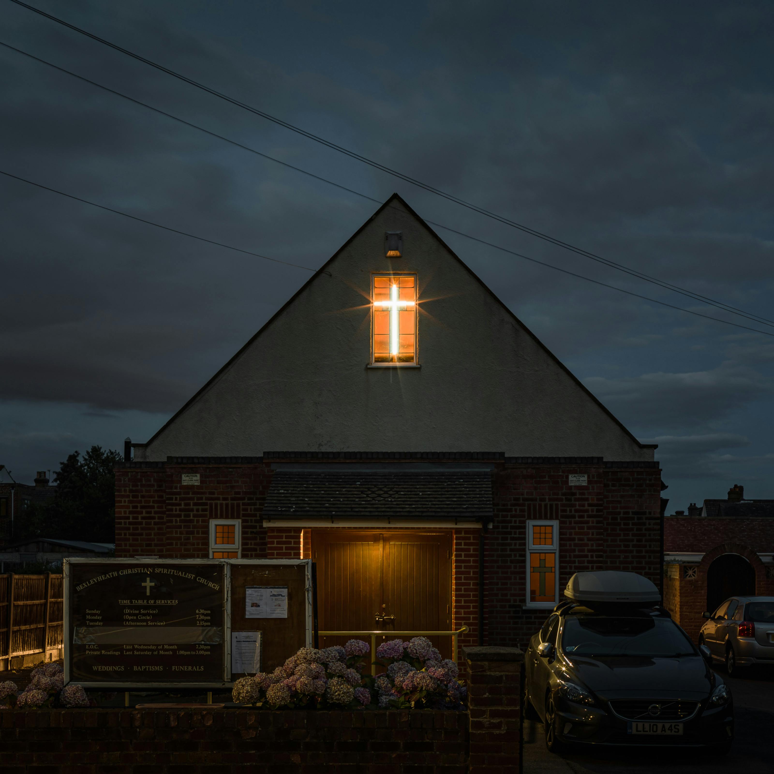 Photograph of Bexleyheath Christian Spiritualist Church at night. The two storey building is set in a residential street, detached but sandwiched between other residential buildings. The second storey shows a brightly lit window with a cross shaped light showing in the window. To the right of the building are two cars.