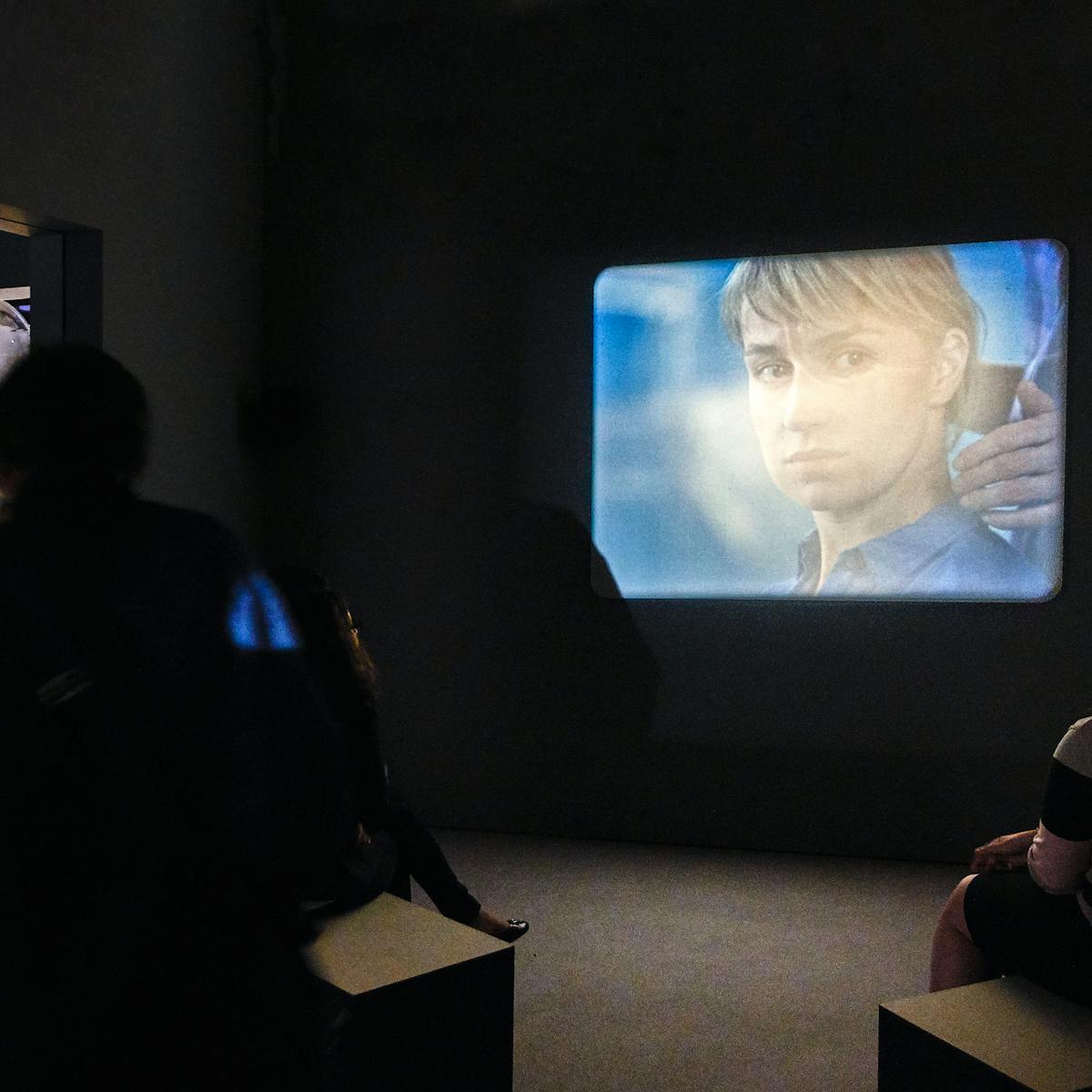 Photograph of film projection showing a woman's face, within a dark gallery setting. In the foreground are visitors sitting on benches watching the film.