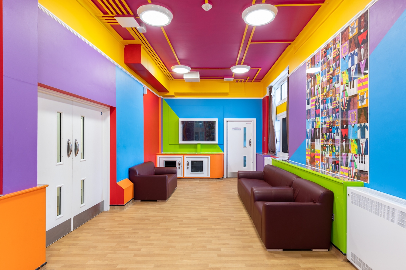 A photograph showing t a long, narrow and brightly coloured room containing a wall-mounted television, and three brown faux leather sofas. The walls are broken up into large purple, blue, red, green and yellow rectangles and triangles. The ceiling is painted purple, with housed wiring painted bright yellow running between the edges of the room and four lights. On the wall behind two sofas, is a square mural made up of lots of smaller squares featuring simple brightly covered graphics including piano keys, clothed torsos, and picture frames. Similar images run along the top of a narrow cupboard below.