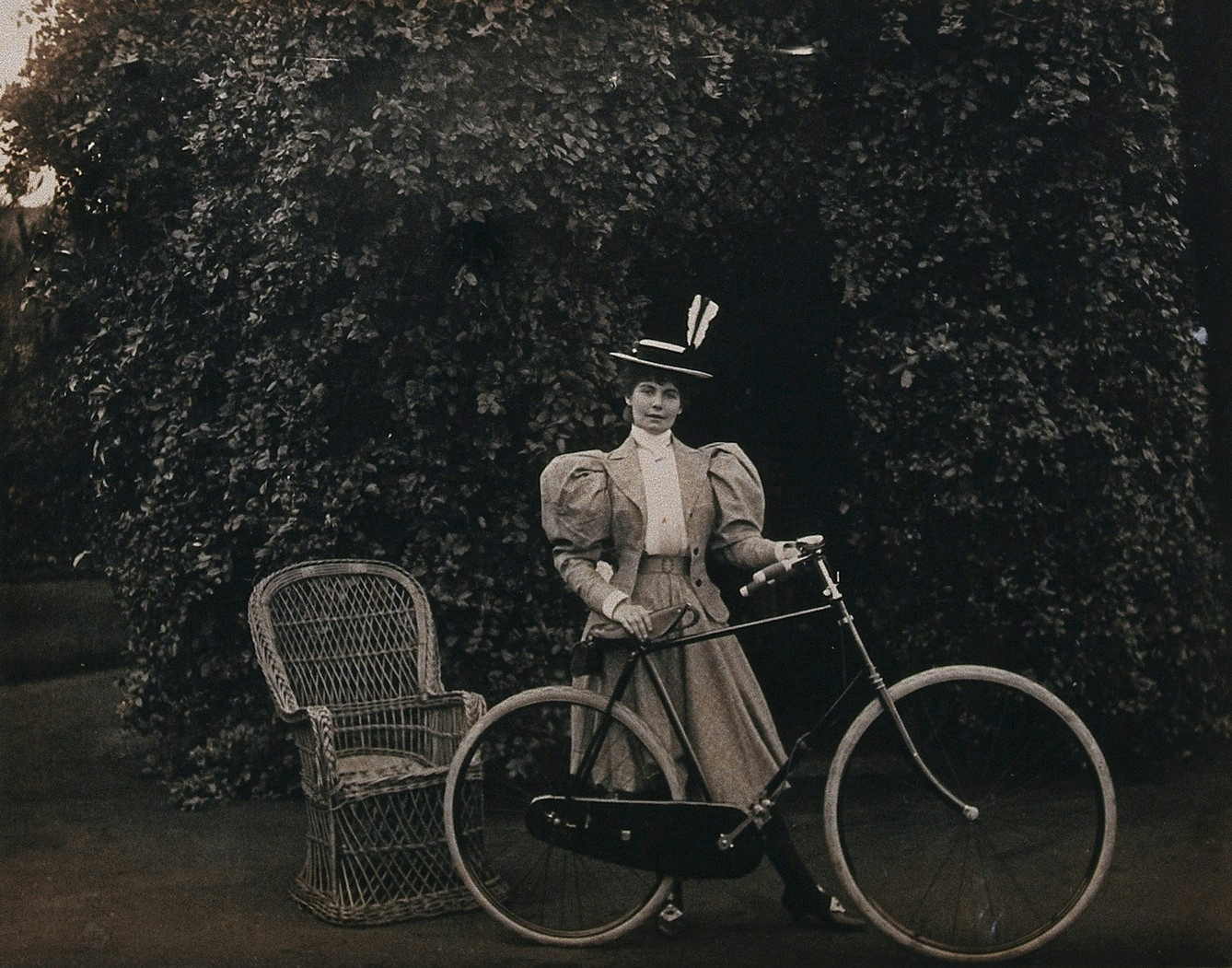 Black and white photo of a woman with a bicycle with bushes in the background. The sleeves on her jacket are puffy and she has two white feathers in her straw boater hat.