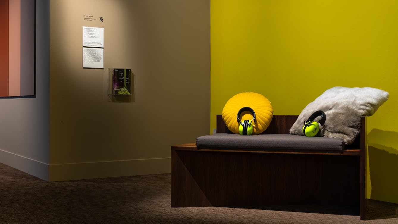Photograph of a corner in a gallery space with green and taupe coloured walls. In the foreground is a gallery seating bench complete with soft cushions and 2 sets of bright yellow/green ear defenders.