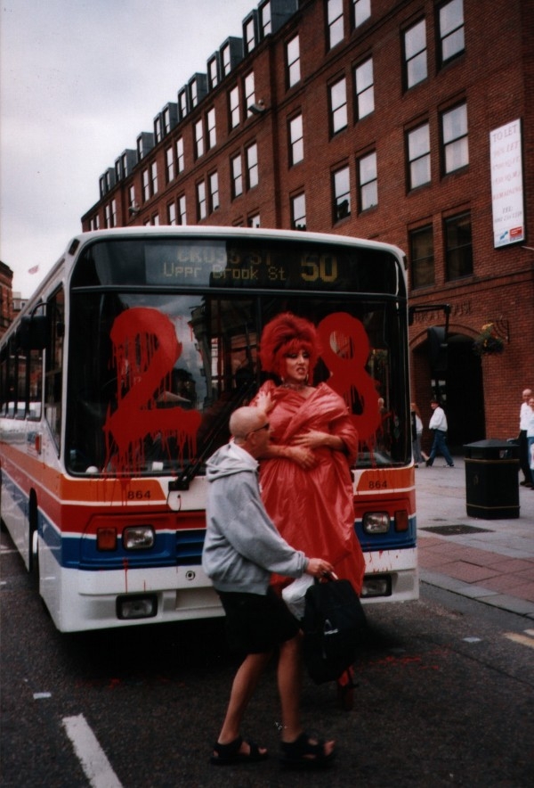 A photograph from July 2000, in which a drag queen wearing red stands in front of a bus. The number "28" has been painted in red, dripping paint onto the windscreen of the bus.