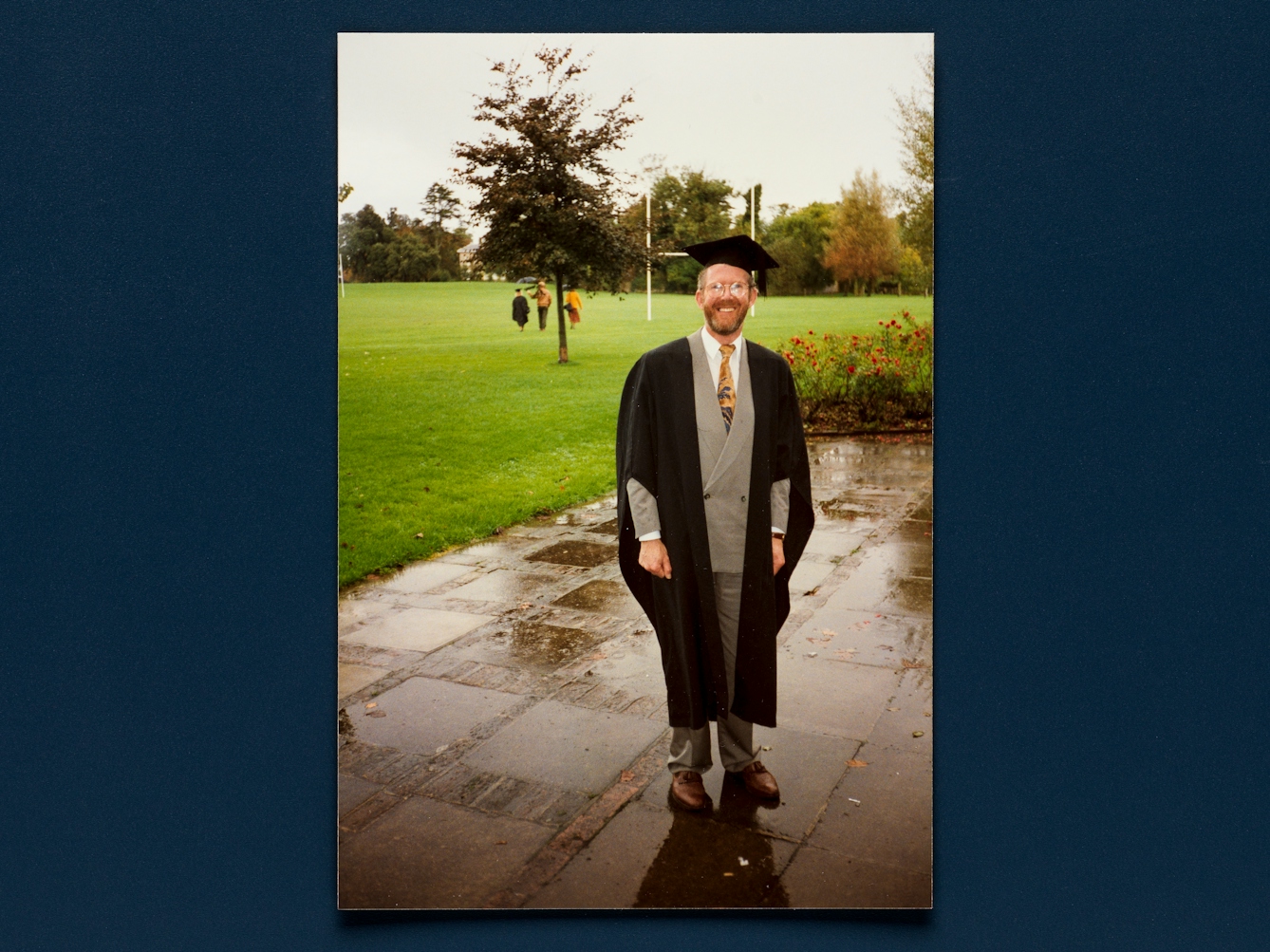Photograph of a print of a colour photo resting on a deep blue background. The photo shows a full-length picture of a man in a grey suit with yellow tie, with glasses and a beard standing outside. He is wearing a graduation gown complete with mortar board.He is smiling to camera. Behind him is a rugby field and he is standing on paved area wet with an earlier rainfall.