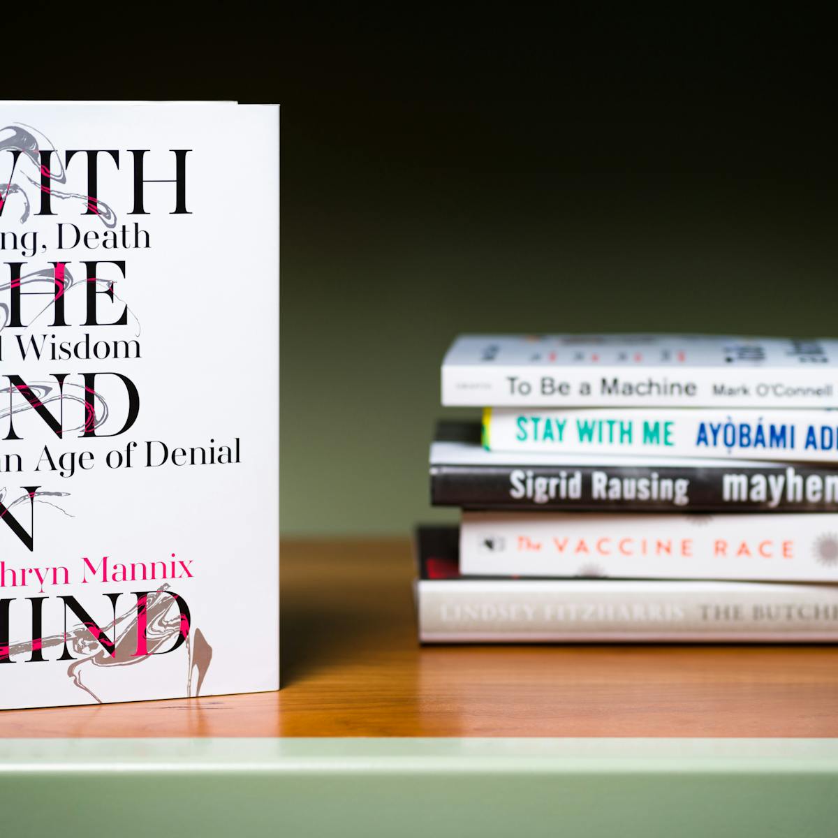 Photograph of a a stack of 5 books lying horizontally on a shelf with another book standing  vertically in the foreground.