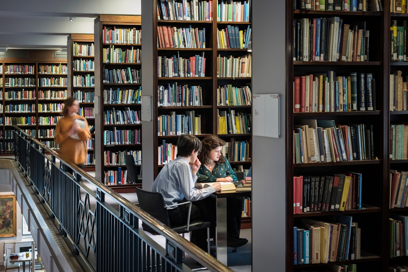 Photograph of two people sat in a library at a table surrounded by floor to ceiling book shelves, filled with different coloured books. Another library user is approaching them with a book in their hands, blurred in the photograph by their movement.