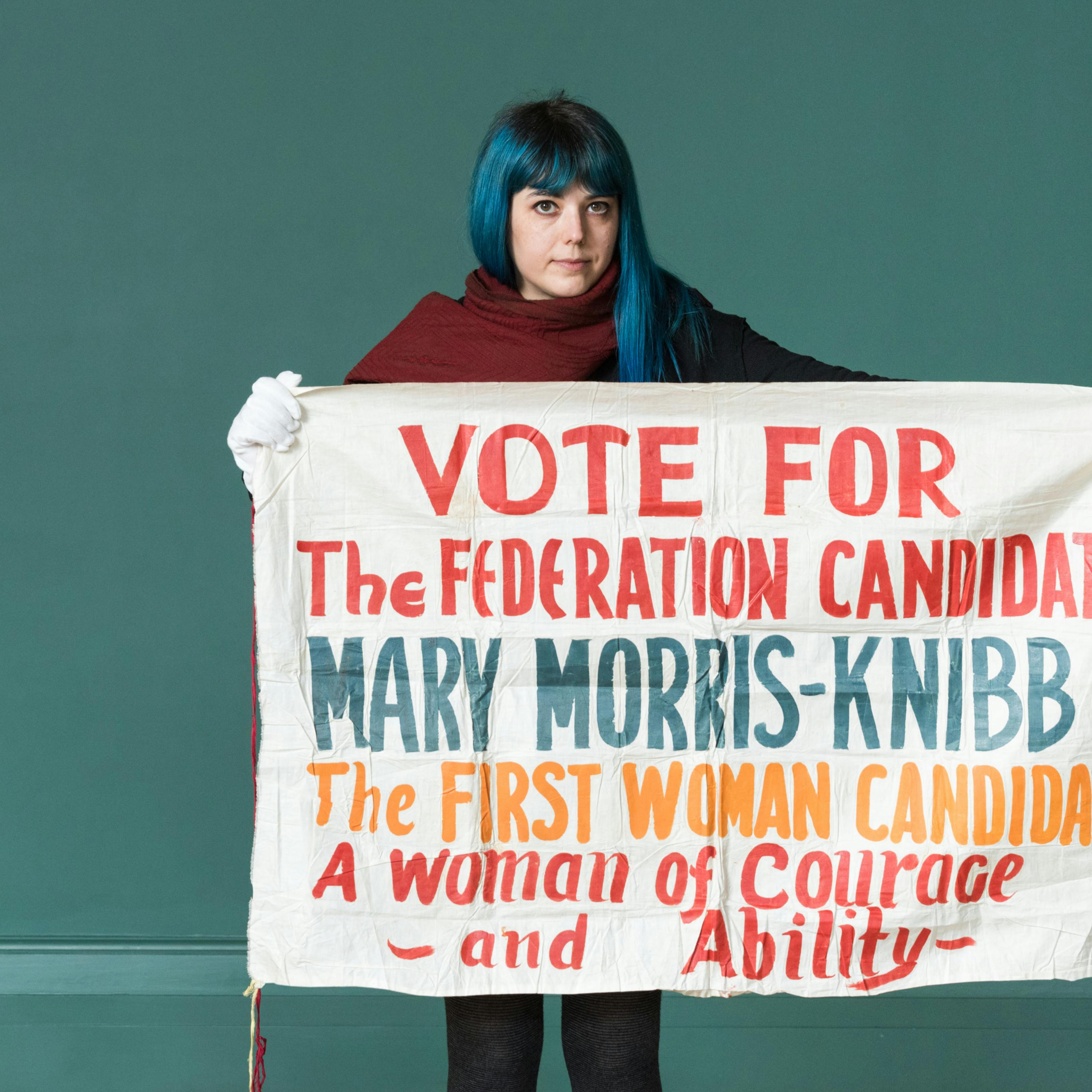 Photograph of a woman holding a 1930s cloth banner with the hand written words 'Vote for the Federation Candidate Mary Morris-Knibb the first woman candidate. A woman of courage and ability'.