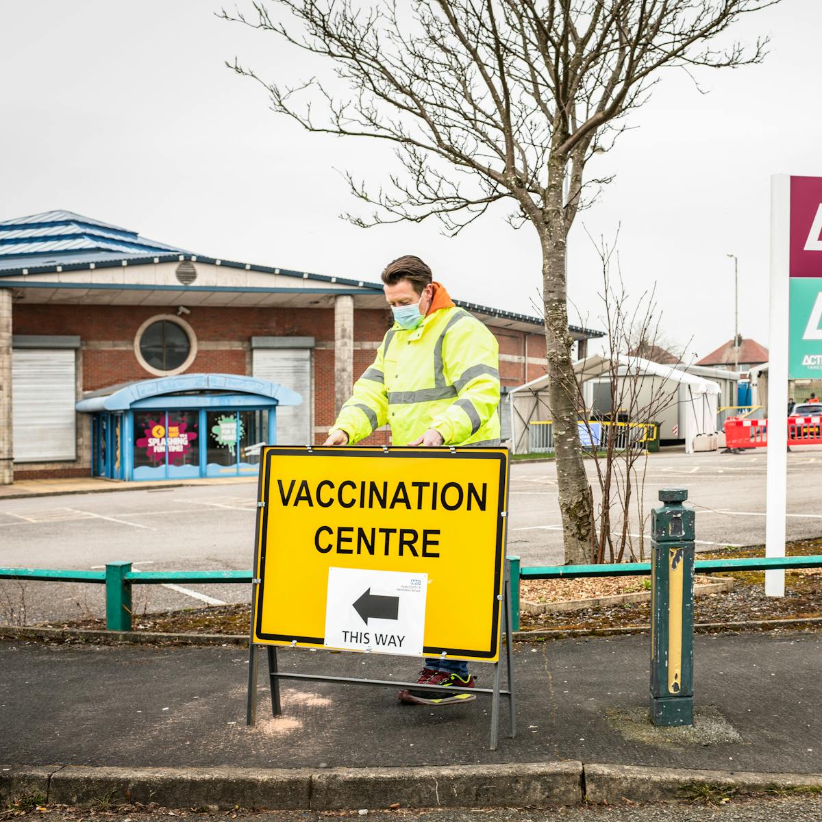 Photograph of the pavement and signage infant of a car park and leisure centre. On the pavement dressed in a hi-vis jacket and a face covering, a man is setting up a freestanding roadsign with the words 'Vaccination Centre this way' written on it. To his right is the large sign to the leisure centre with the words 'Hyde' and 'Hyde Leisure Pool' displayed. In the background is the carpark where temporary marquees have been erected.