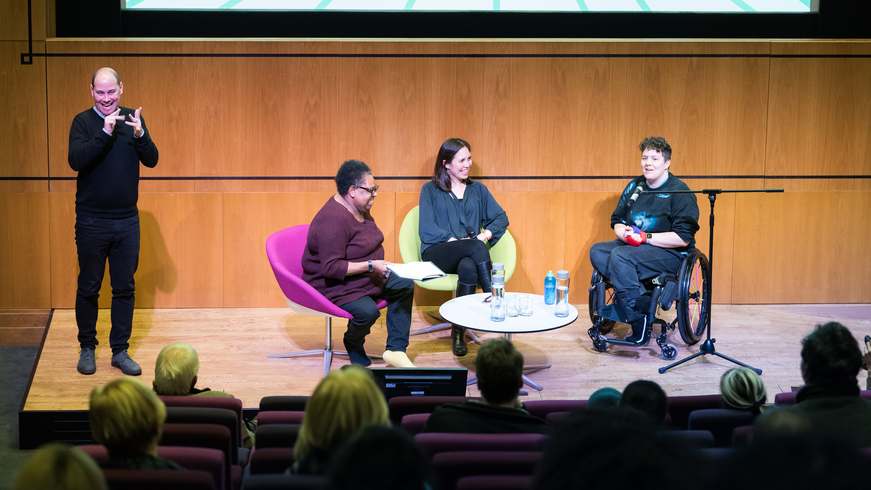 Jess Thom speaking to an audience at Wellcome Collection. Next to her is a panel of speakers and a BSL Interpreter.