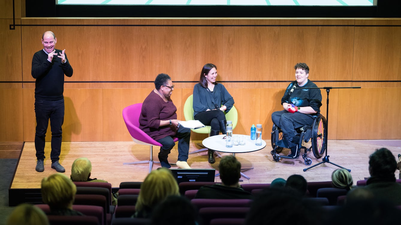 Jess Thom speaking to an audience at Wellcome Collection. Next to her is a panel of speakers and a BSL Interpreter.