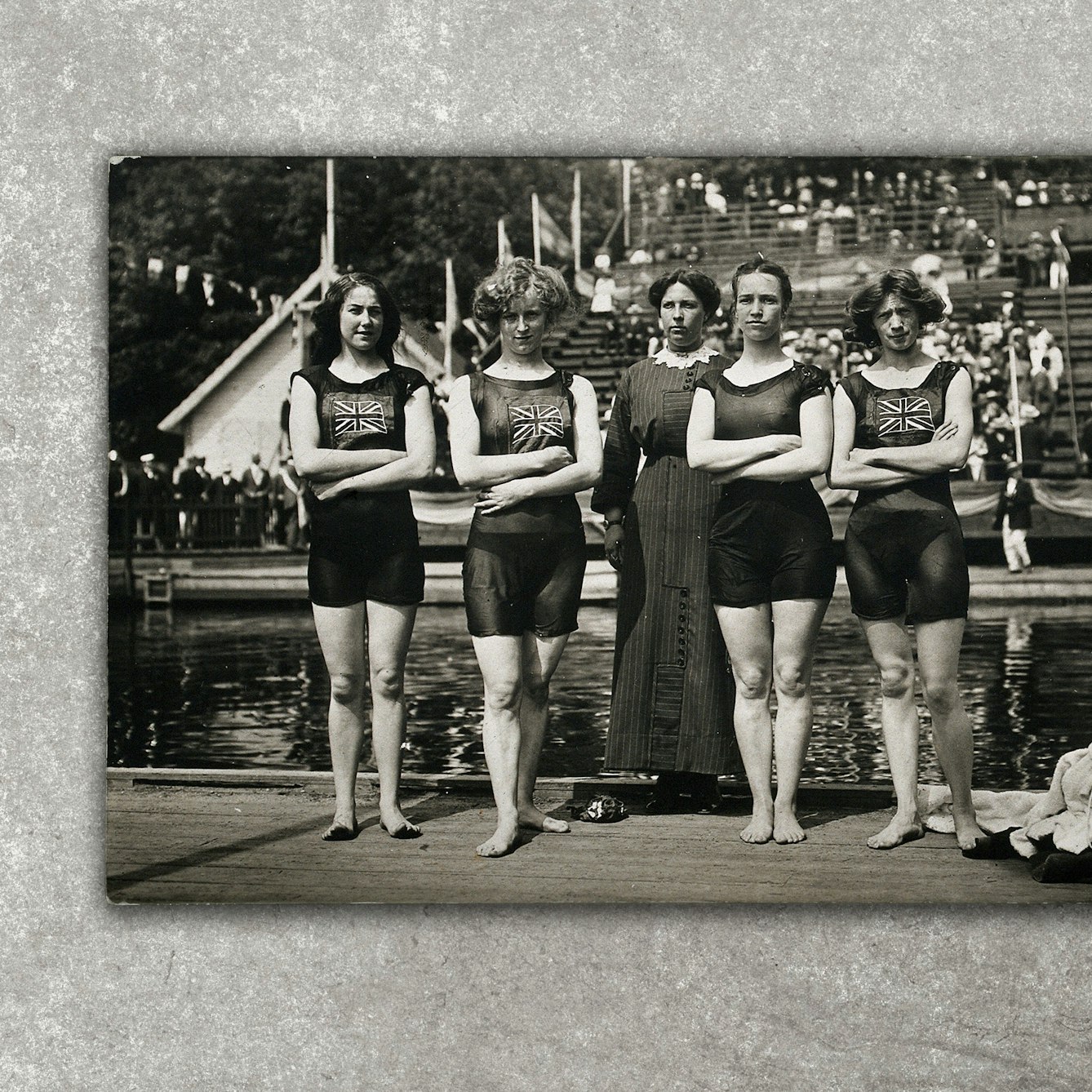 Photograph of a black and white photographic postcard showing the victorious English 400m women's relay swimming team at the Stockholm Olympic Games in 1912. It shows 4 female swimmers wearing swimming costumes with Union jack flags on the front, standing in a line beside an open air pool. In the middle of the four swimmer is another lady who is fully clothed. The postcard has been photographed against a grey concrete background.