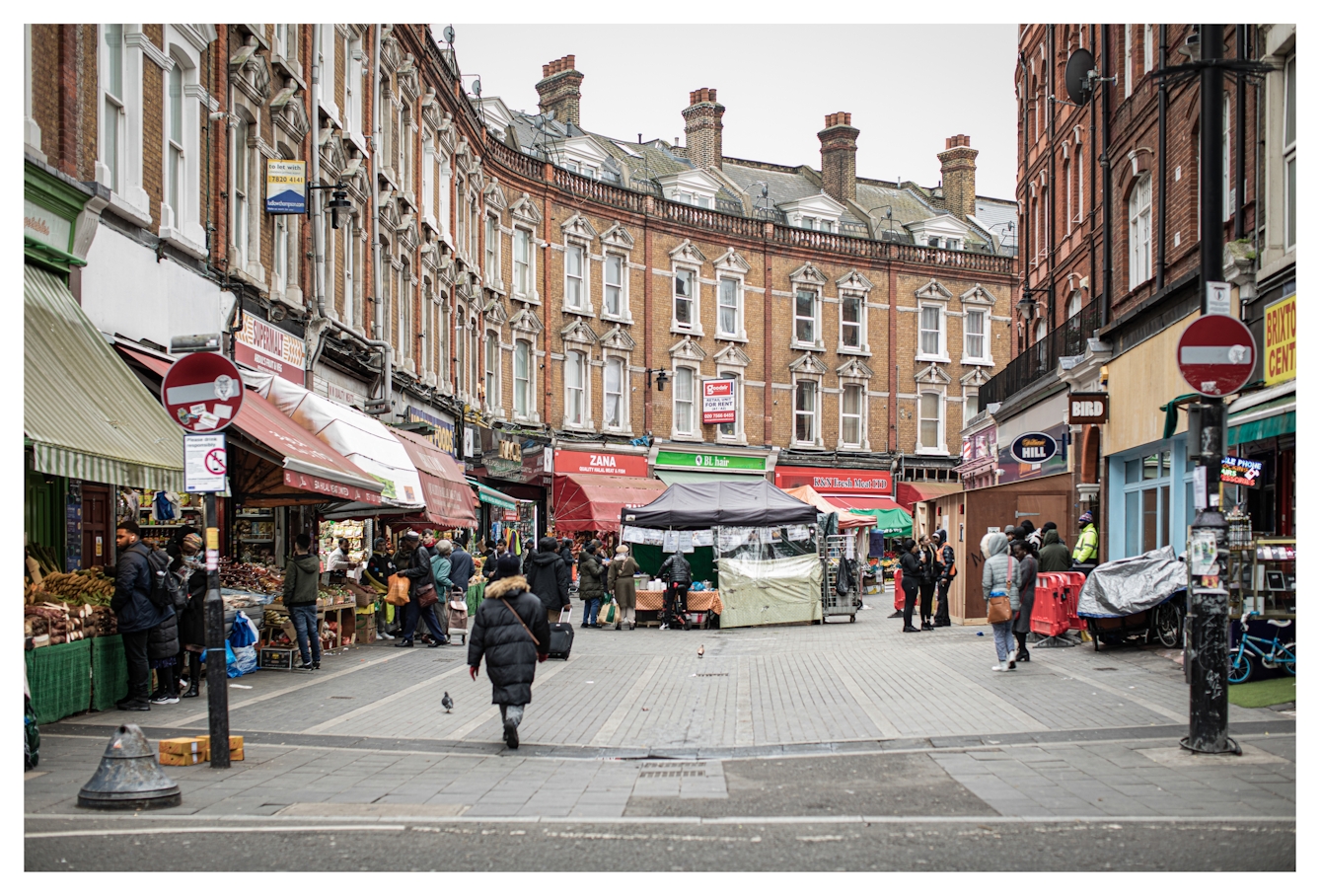 Photograph of a South London street scene in Brixton, showing a pedestrian road with market stalls set up in the middle. The street is flanked by tall victorian red brick buildings. The ground floors are all shops, some with awnings lowered. People are milling about around the market stalls, carrying bags. In the foreground to the left and right are two posts with circular red and white no entry road signs.