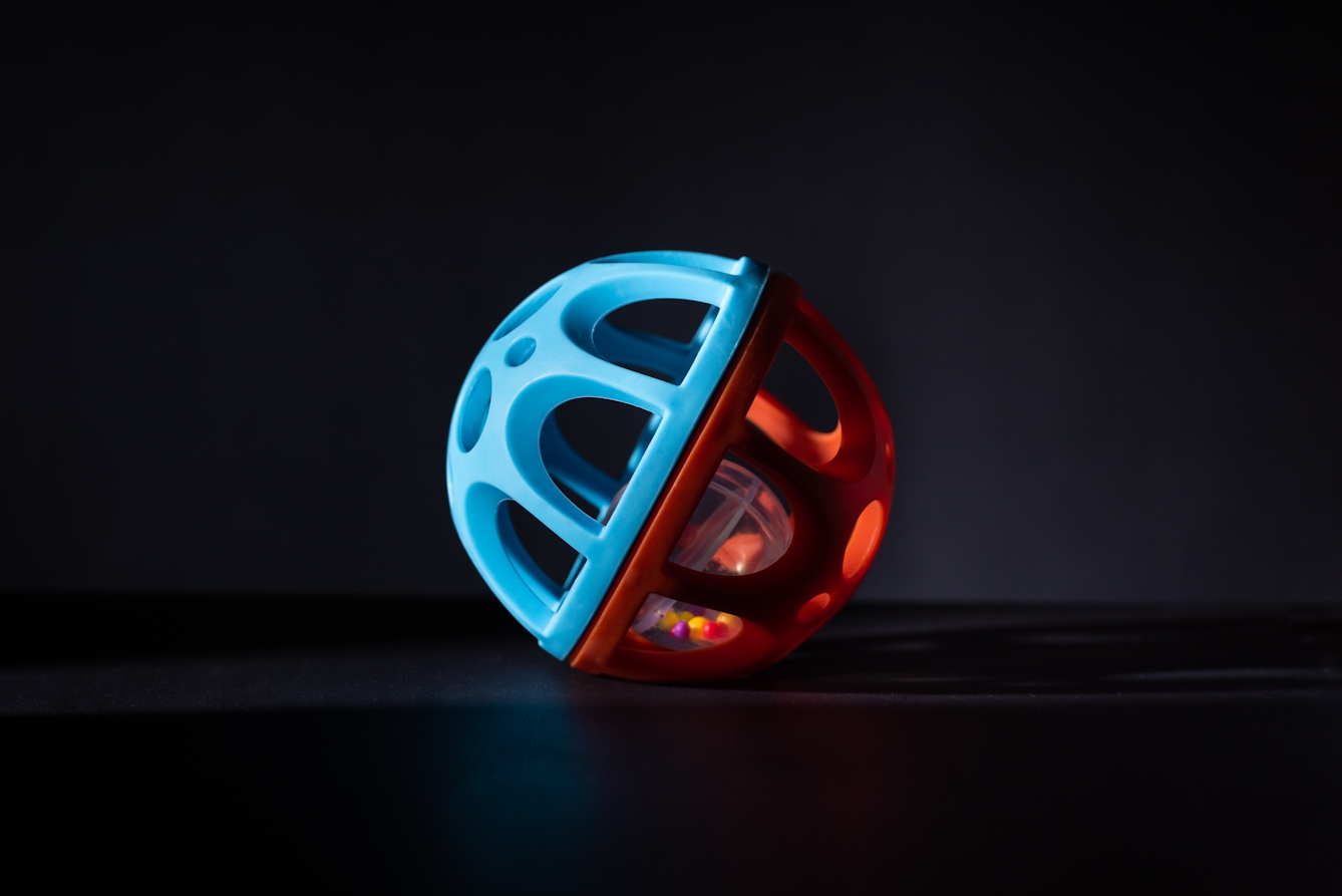 A photograph of rattle ball toy against a black background. The ball is half orange and half blue appears so that one half is in dark shadow and silhouette.
