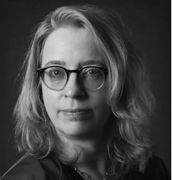 Black and white photograph of the head of a middle-aged woman with shoulder-length blonde hair and glasses. The woman is looking directly at the viewer with a slight smile.