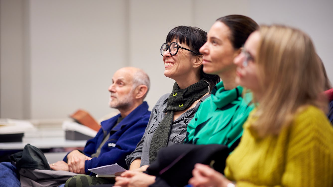 Photograph of four people seated in a row at a talk. They are looking up to listen to a speaker who is out of frame.