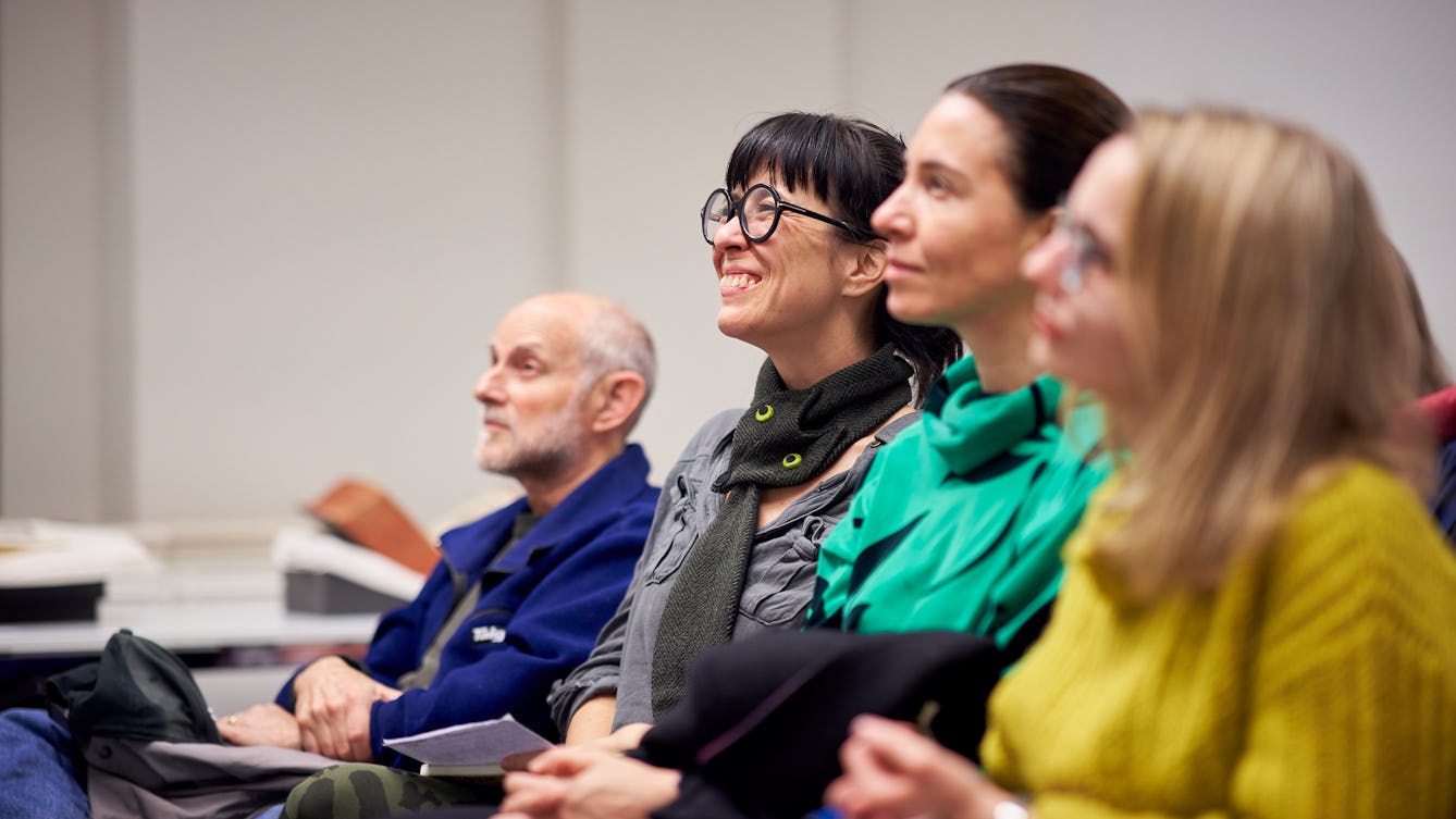 Photograph of four people seated in a row at a talk. They are looking up to listen to a speaker who is out of frame.