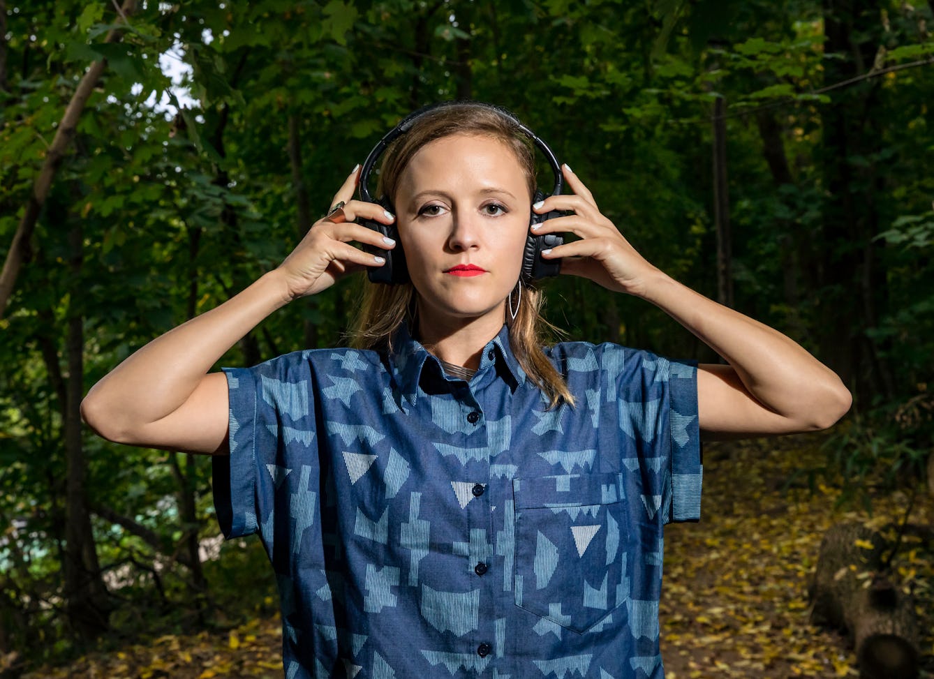 A photograph of a woman with long brown hair holding a pair of headphones to her ears, surrounded by trees.