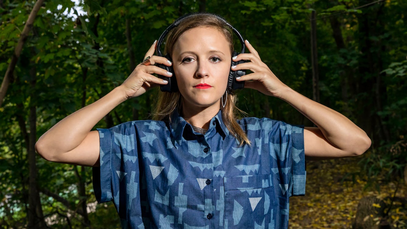 A photograph of a woman with long brown hair holding a pair of headphones to her ears, surrounded by trees.