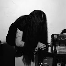 Photograph of a woman crouching, holding an object  in her hand and looking at it, her face obscured by long dark hair.